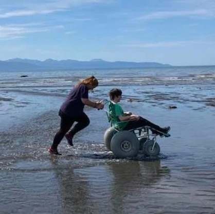 A woman pushing a boy in a wheelchair across the beach in shallow water.